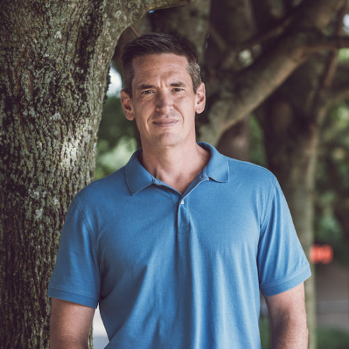 Brunette man in blue shirt standing outside before some trees.