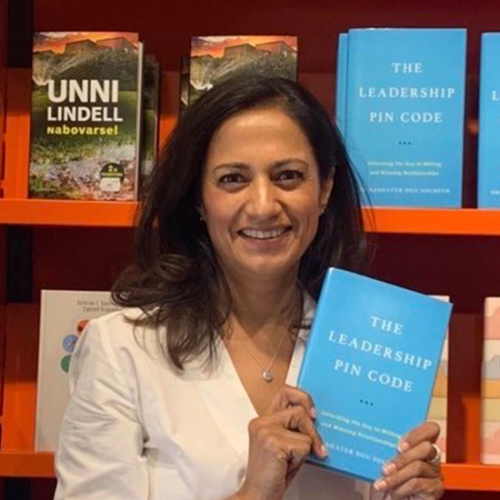 Brunette woman in white top stands in front of book shelves holding up a blue book.