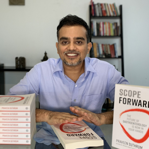 Indian man in blue shirt sits at table with stack of books.